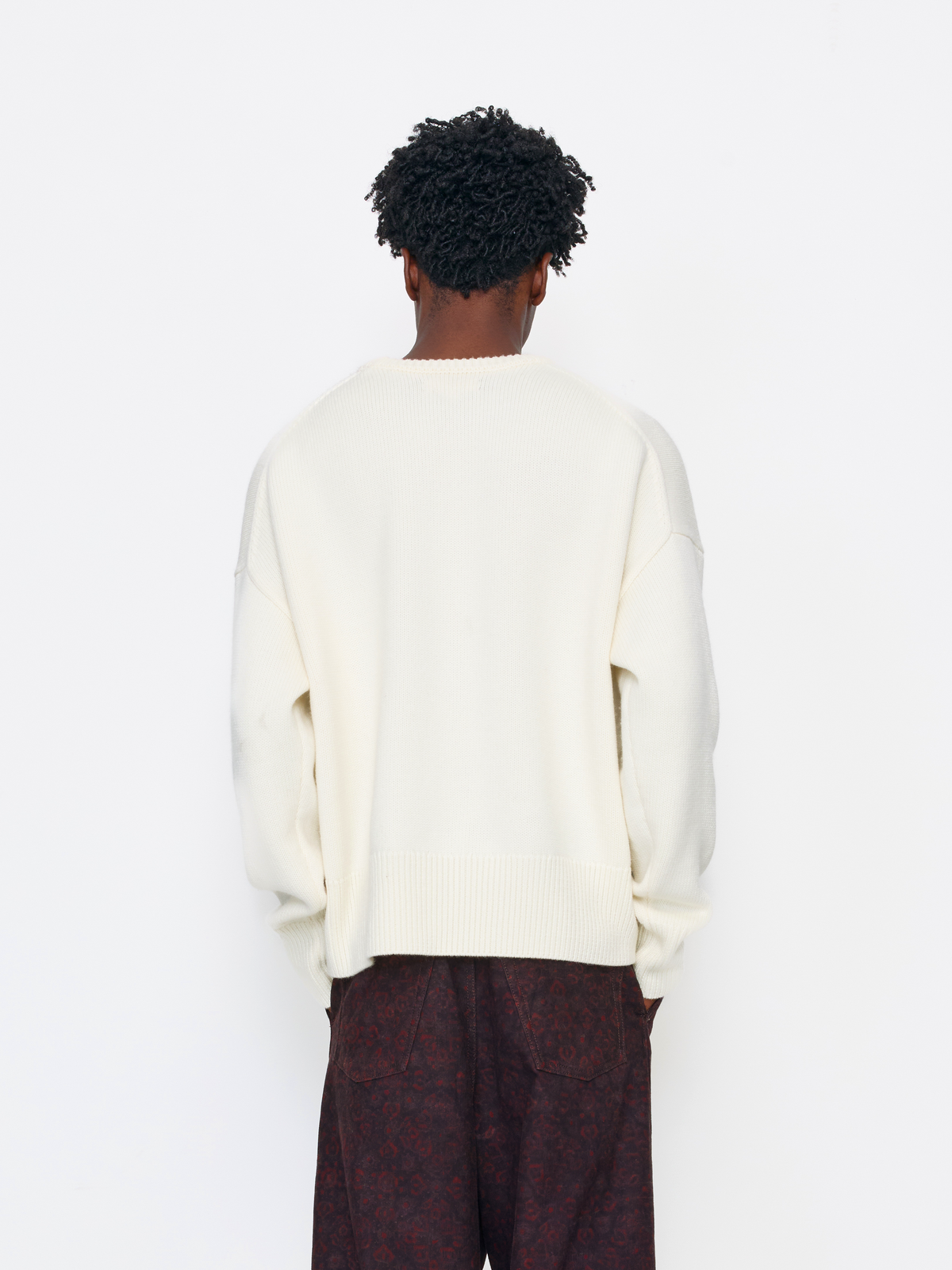 A person shown from the back wearing an oversized cream knit sweater and dark patterned trousers against a plain white studio background.