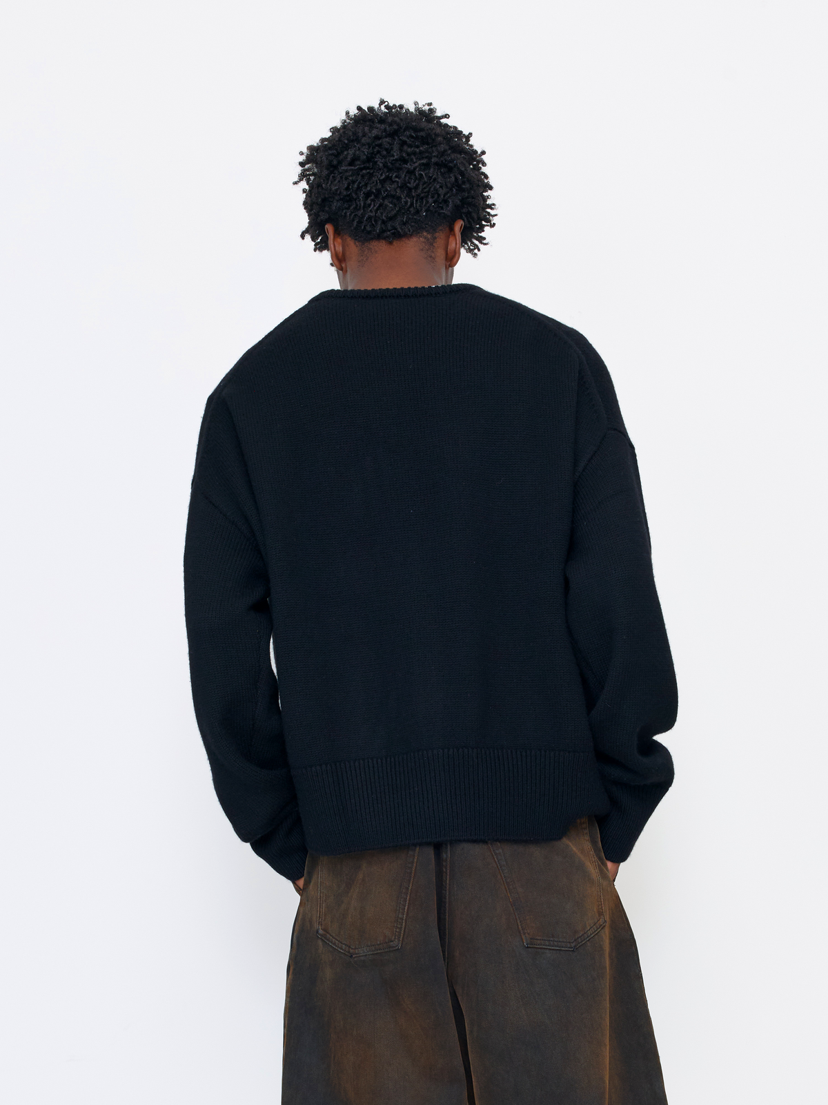 A person shown from the back wearing a black knit sweater and brown wide-leg pants against a plain white studio background.