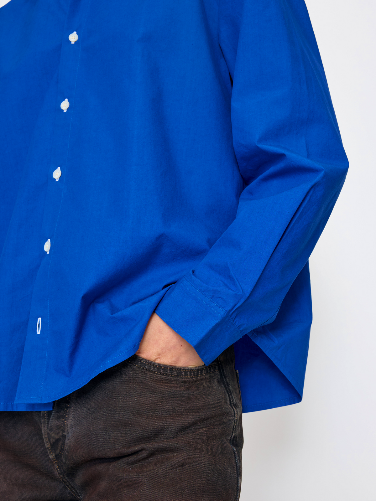 A close-up of a person wearing a bright royal blue long-sleeve button-up shirt tucked into dark brown or black corduroy pants, with one hand in the pocket.