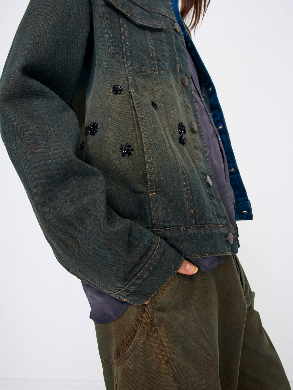 A close-up of a person wearing a dark washed denim jacket with black floral appliqués, layered over a purple shirt and paired with olive green pants, against a plain white background.