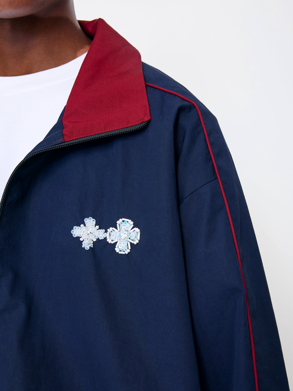 Close-up of a navy blue track jacket with a red collar, red piping, and two silver rhinestone cross brooch pins on the chest, worn over a white t-shirt.