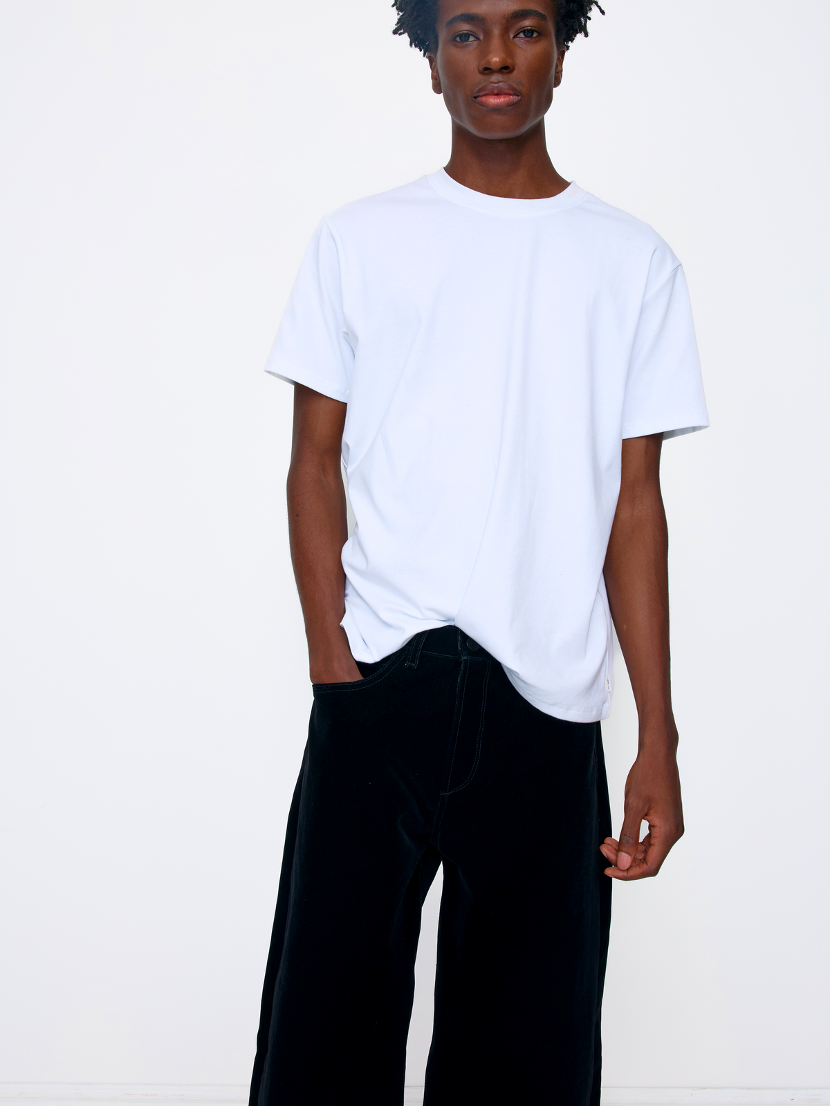 A model wearing a plain white short-sleeve crewneck t-shirt and black wide-leg velvet pants, standing against a clean white studio background.