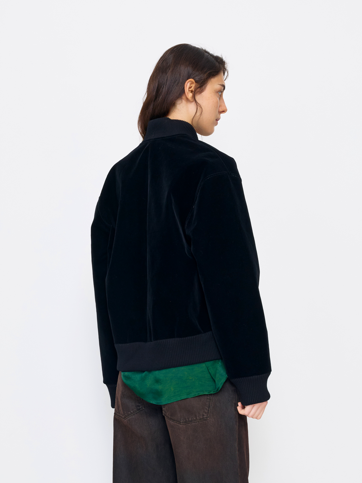 A model shown from the back wearing a black velvet jacket with ribbed trim, layered over a green shirt and dark brown pants, against a plain white studio background.