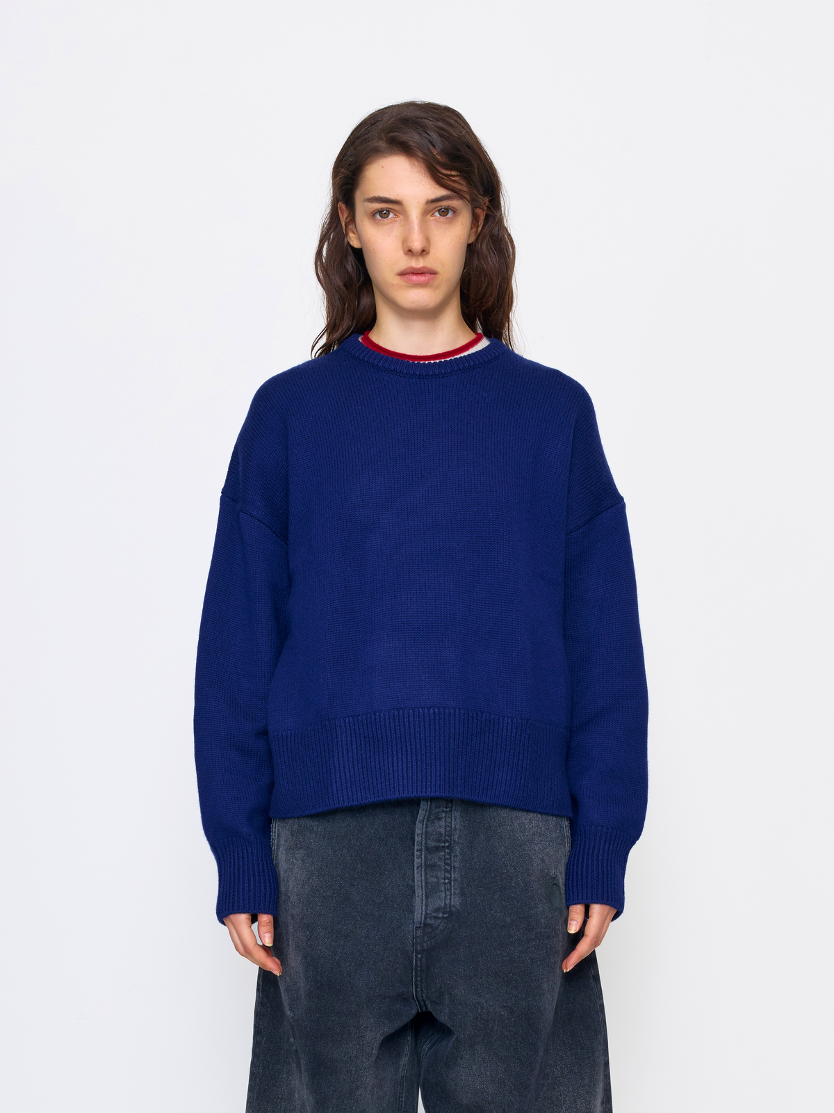 A model wearing a dark blue crewneck sweater with a red and white striped collar, paired with dark gray jeans, against a plain white studio background.