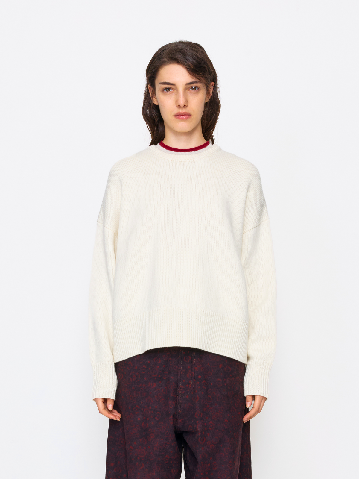 A model wearing an oversized cream-colored knit sweater with a contrasting dark red crew neck, paired with dark patterned trousers, shot against a solid white studio background.