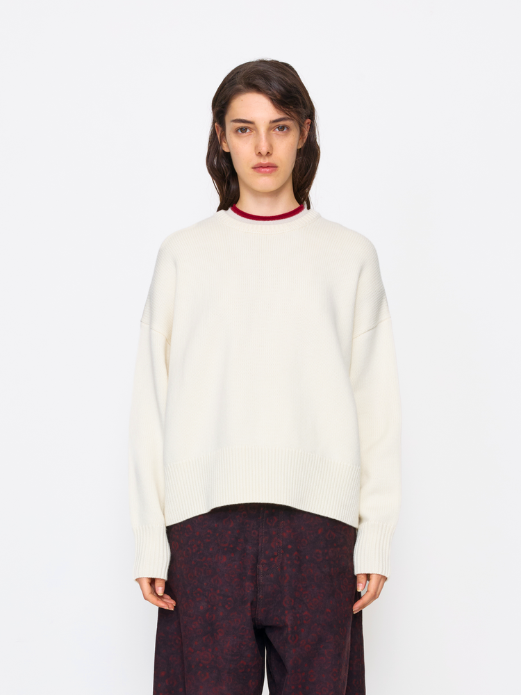 A model wearing an oversized cream-colored knit sweater with a contrasting dark red crew neck, paired with dark patterned trousers, shot against a solid white studio background.