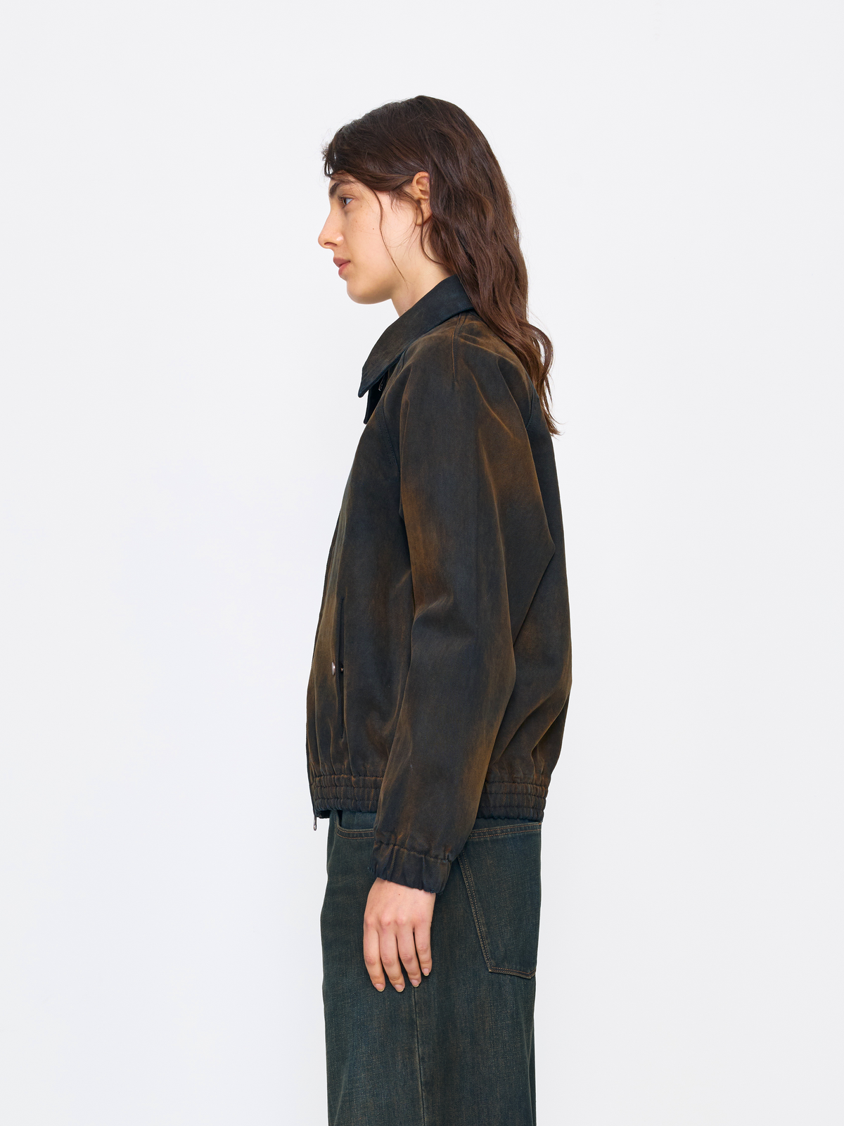 A model shown in profile wearing a dark brown and black acid-washed jacket with dark blue jeans, set against a plain white studio background.