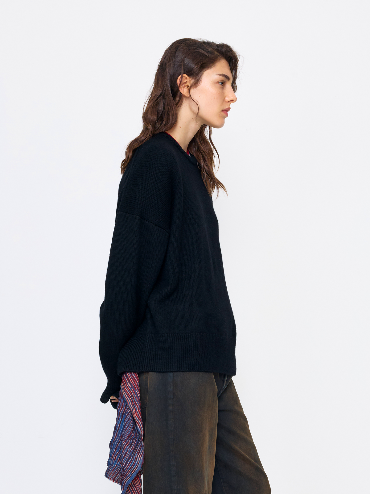 A woman shown in side profile wearing a black knit sweater and dark brown pants, standing against a plain light gray studio background.