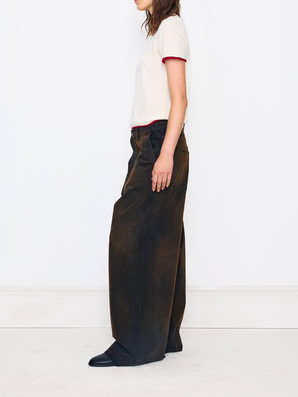 A model is shown from the side wearing a cream short-sleeve t-shirt with red trim and very wide-leg dark brown trousers, paired with black shoes, against a clean white studio background.