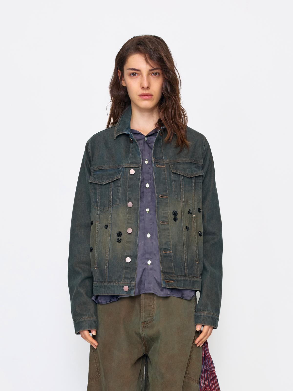 A model wearing a dark washed denim jacket with distressed details, layered over a purple button-up shirt and olive green pants, set against a plain light gray background.