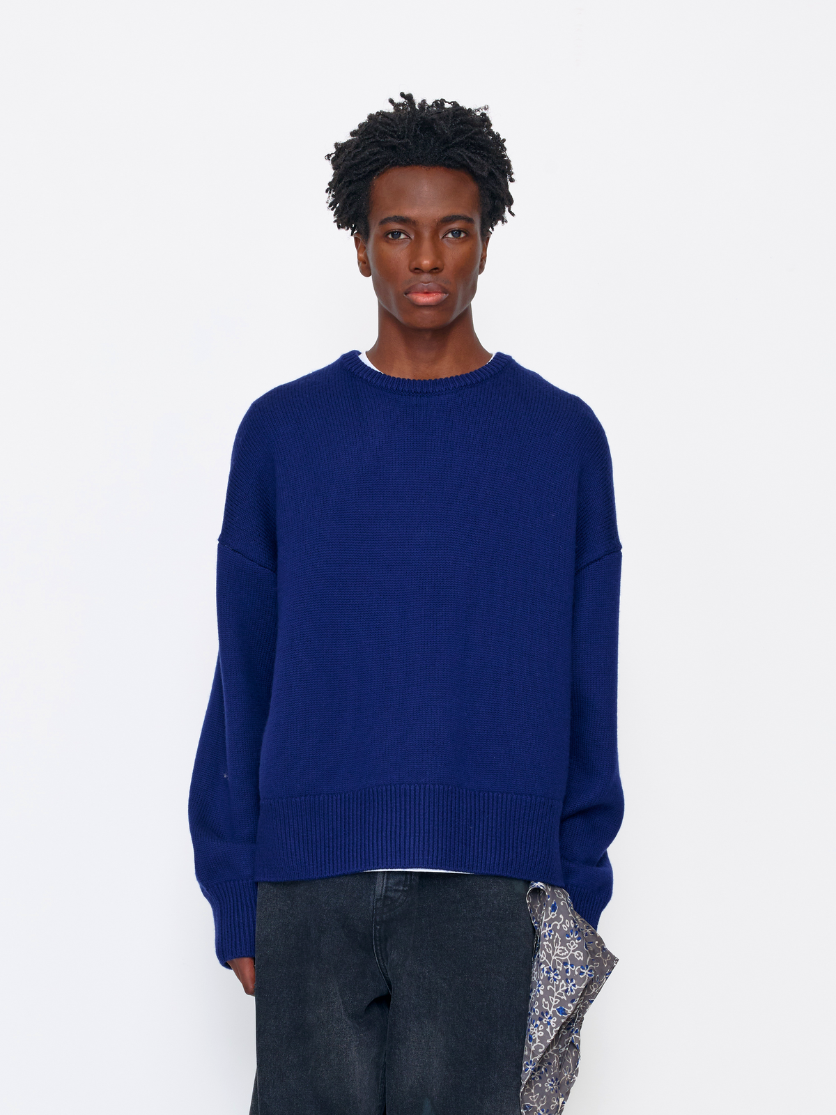 A model wearing a vibrant royal blue crewneck sweater, paired with dark gray jeans and a patterned clutch, shot against a solid white studio background.