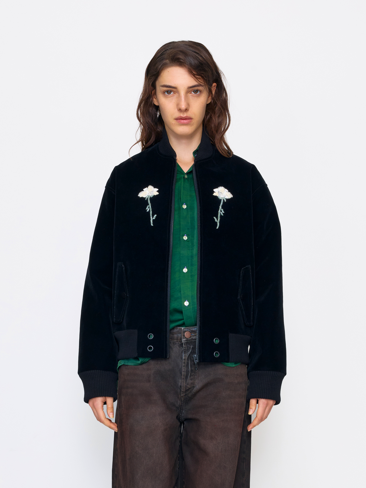 A model wearing a black velvet bomber jacket with white embroidered flowers on the chest, layered over a green button-up shirt and paired with brown pants, shot against a plain white studio background.