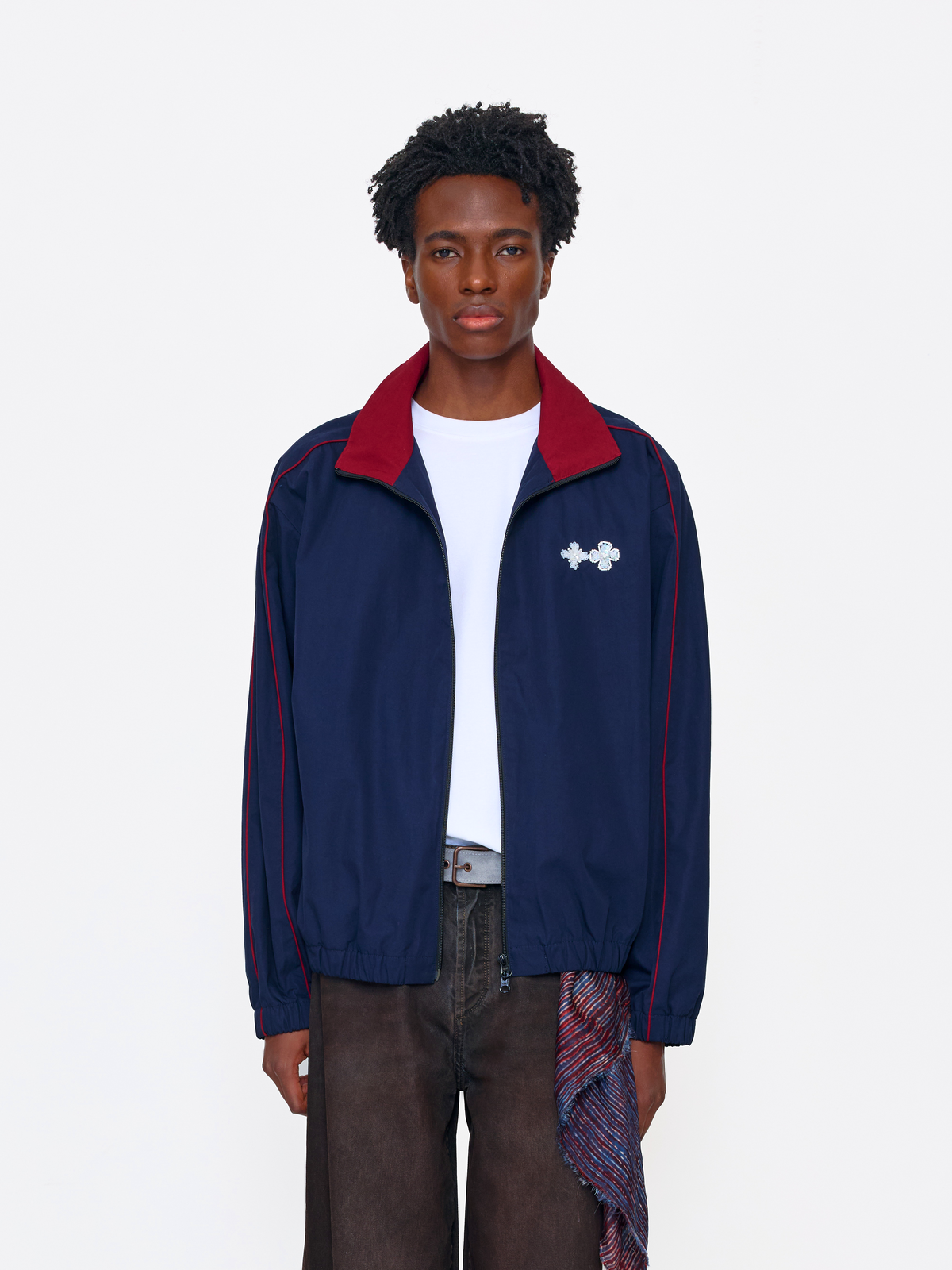 A model wearing a navy blue track jacket with a red collar and red piping, featuring a white Under Armour logo on the chest, styled over a white t-shirt and dark brown pants.