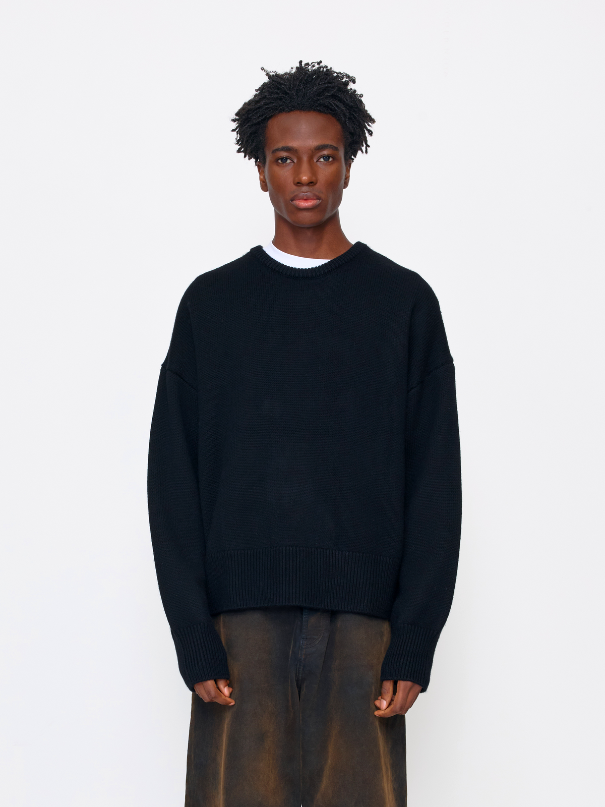 A model wearing a black knit crewneck sweater over a white undershirt and brown trousers, standing against a plain white studio background.