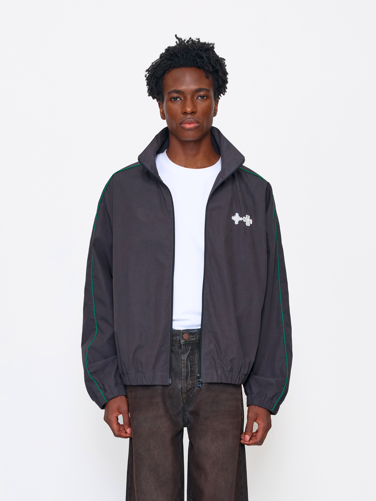 A model wearing a dark gray zip-up jacket with green piping and a white Under Armour logo on the chest, layered over a white t-shirt and dark brown pants, against a plain white studio background.