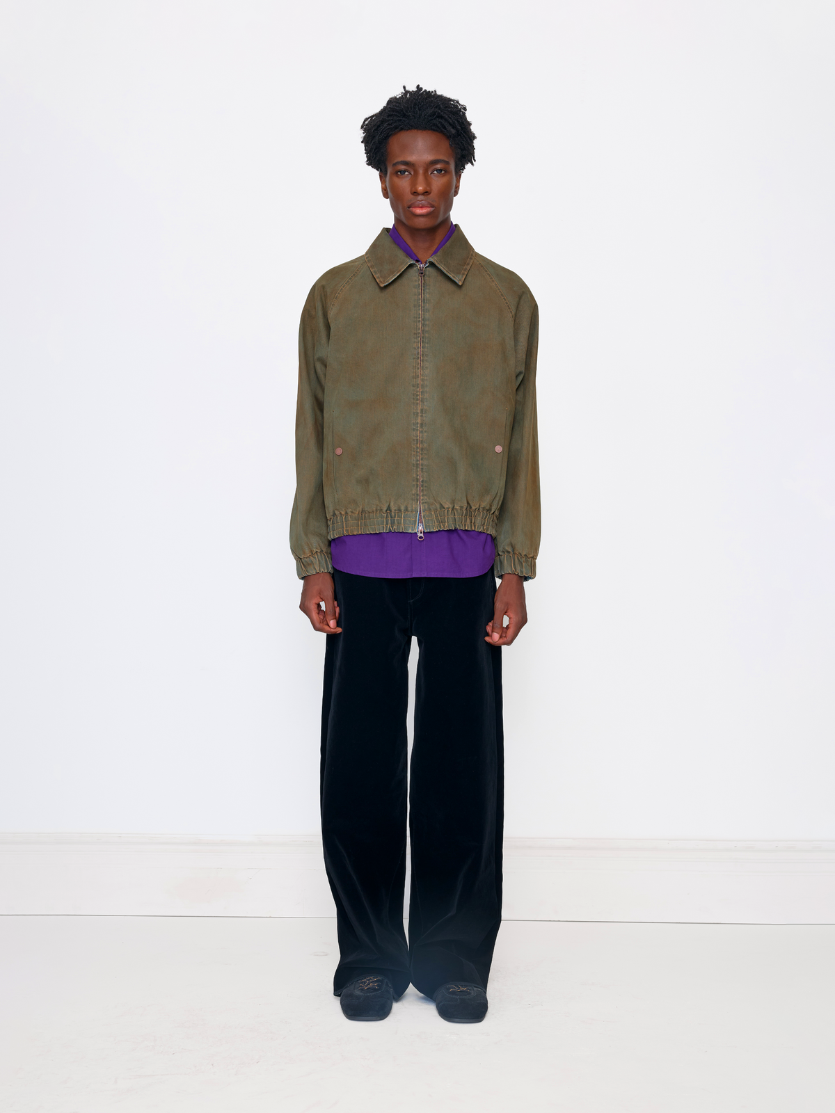 A model stands against a plain white studio background wearing an olive green cropped jacket over a purple shirt, paired with black velvet trousers and dark loafers.
