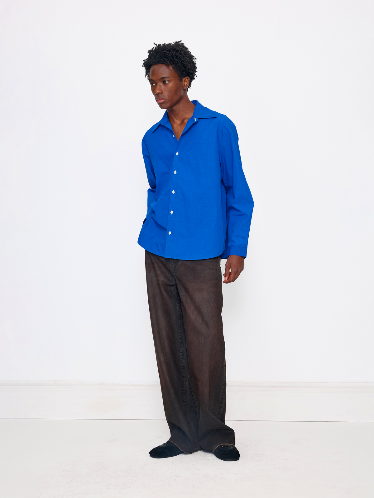 A model stands in a studio wearing a bright royal blue long-sleeve button-up shirt and loose dark brown wide-leg trousers.