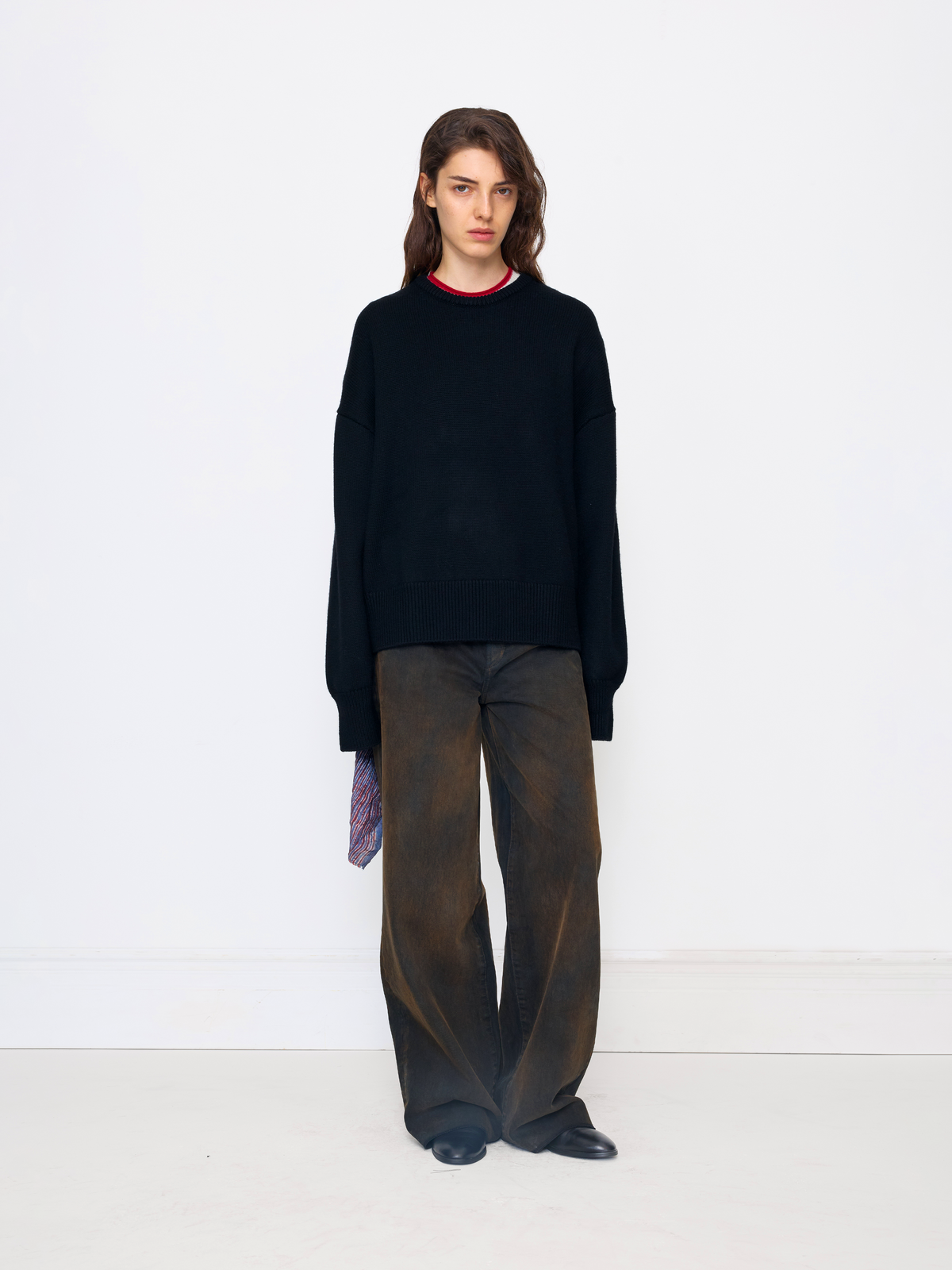 A model stands against a plain white studio wall wearing an oversized black knit sweater with a thin red collar trim, loose dark brown wide-leg trousers, and black shoes.