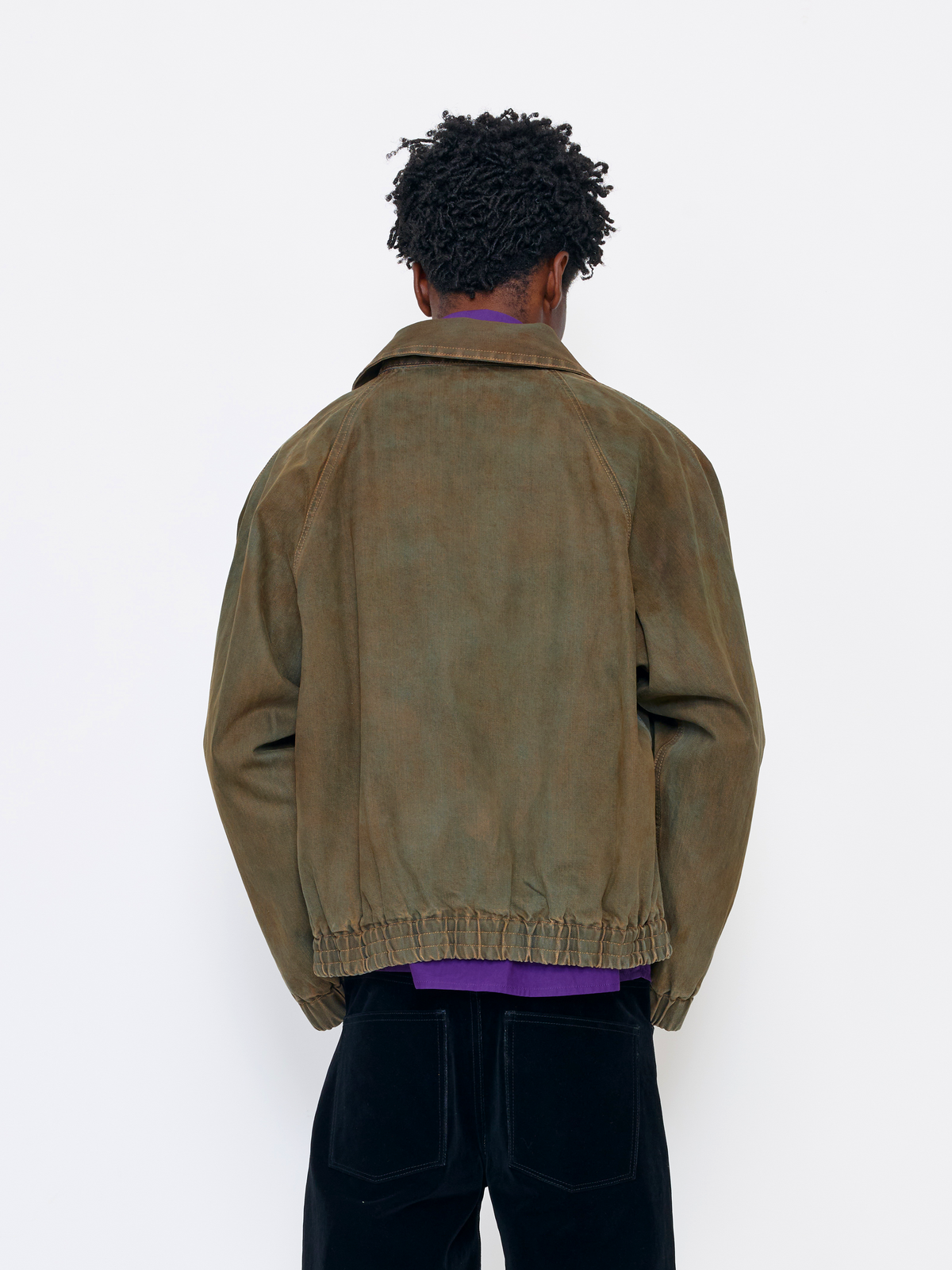 A person shown from the back wearing an oversized brown suede or washed leather jacket over a purple shirt and black pants, standing against a plain white wall.