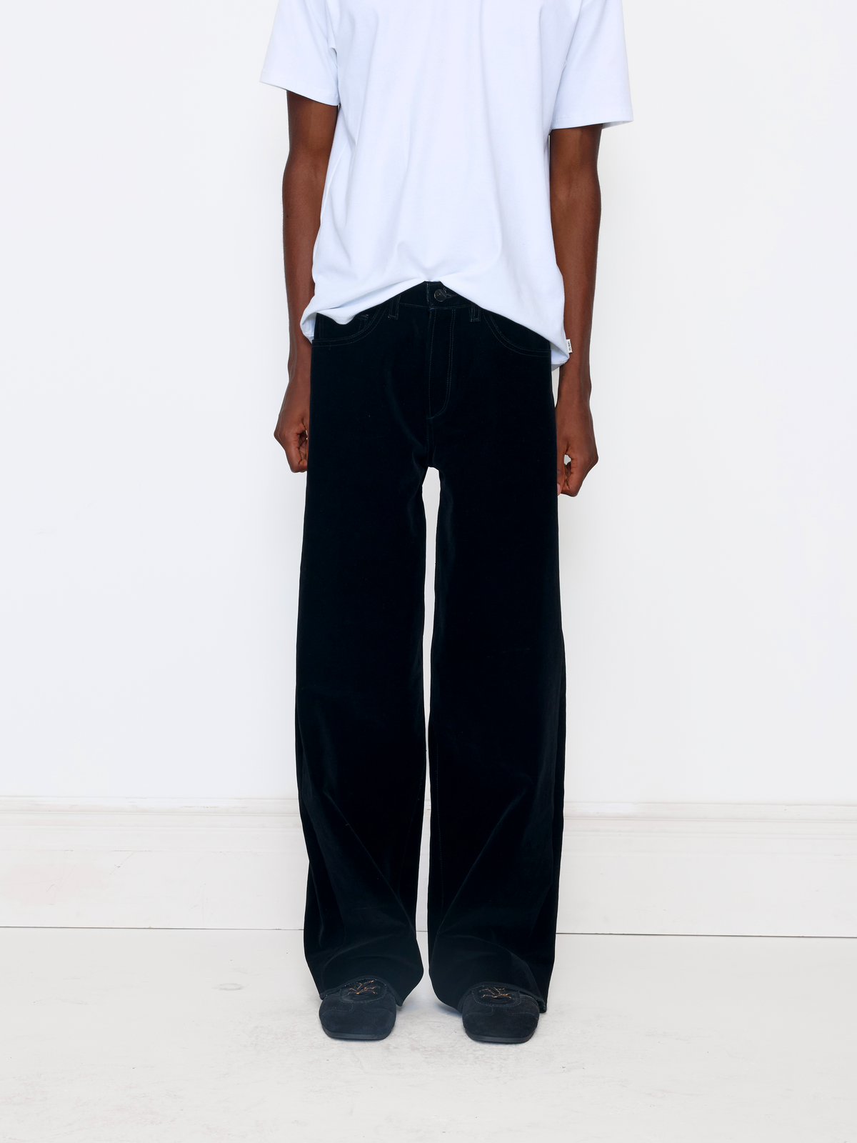 A model wearing black wide-leg velvet trousers, paired with a white t-shirt and black shoes, standing against a plain white studio background.
