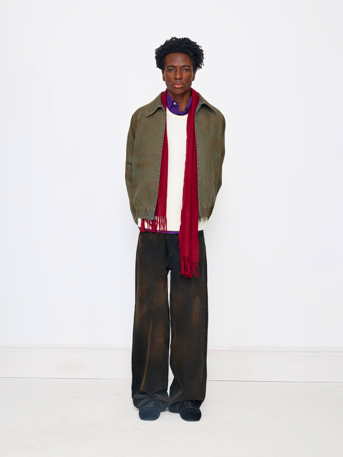 A model stands against a plain white studio background wearing an olive green jacket, a white sweater, a red scarf, dark brown wide-leg trousers, and dark blue shoes.