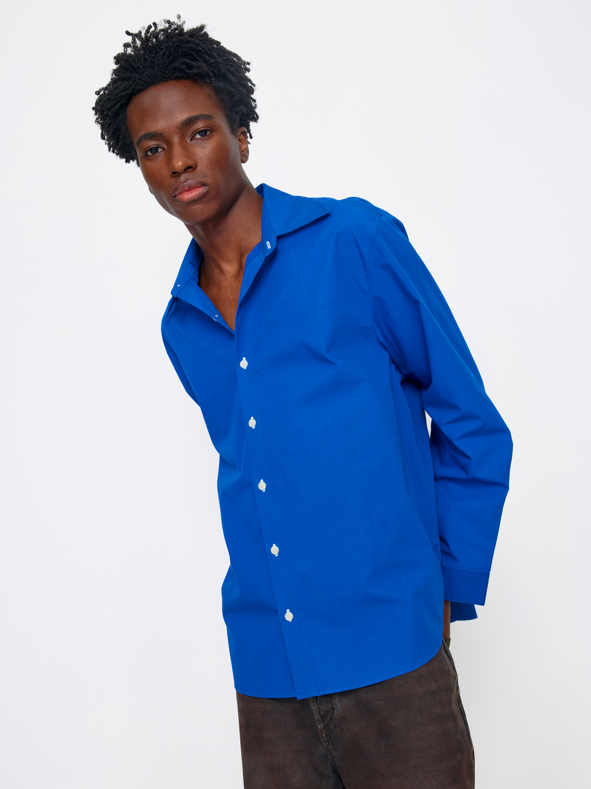 A man wearing a bright royal blue long-sleeve button-up shirt and dark brown pants, photographed against a clean white studio background.