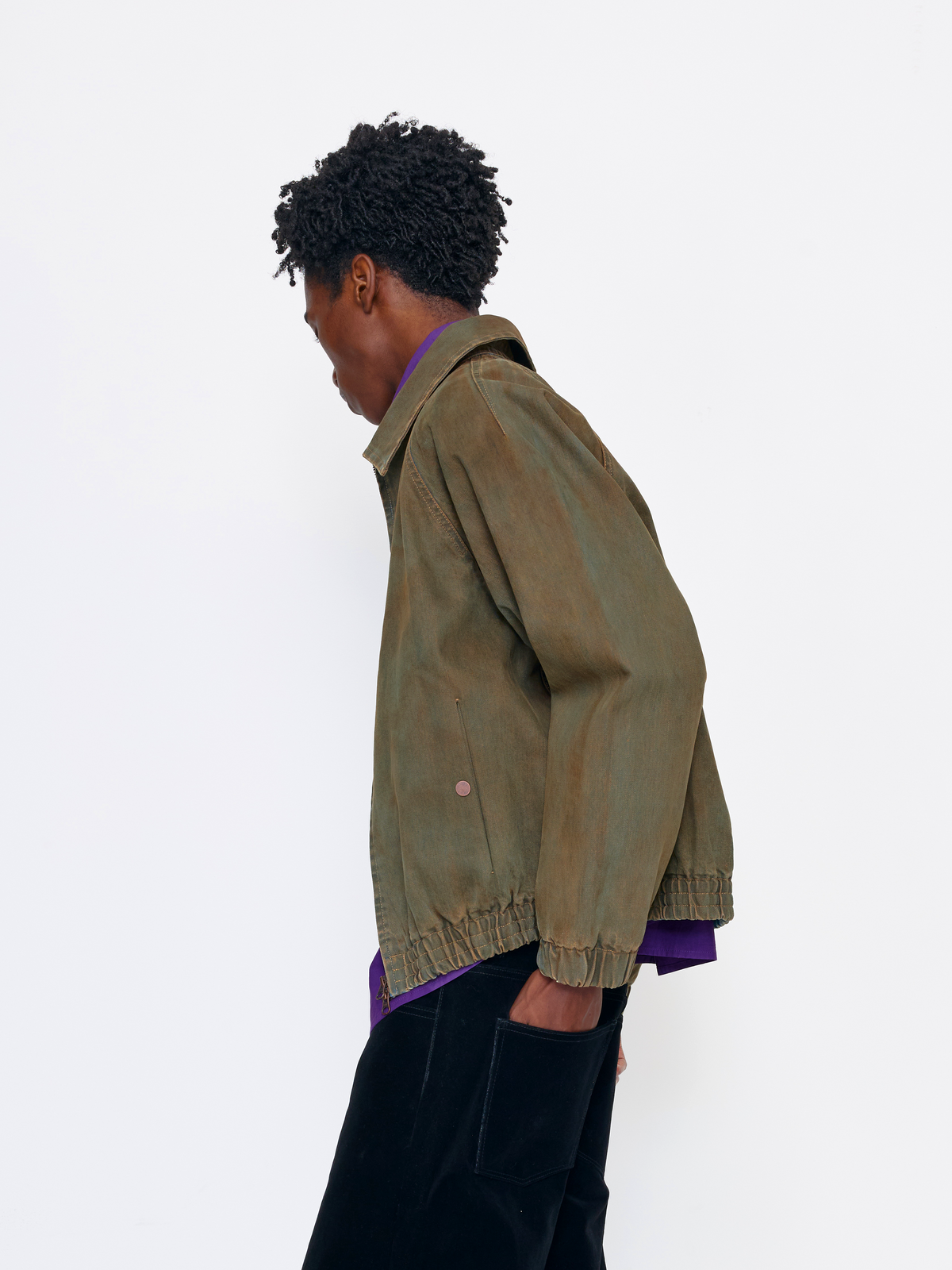 A model shown from a side-back angle wearing an olive green canvas utility jacket over a purple shirt and black pants, set against a plain white studio background.