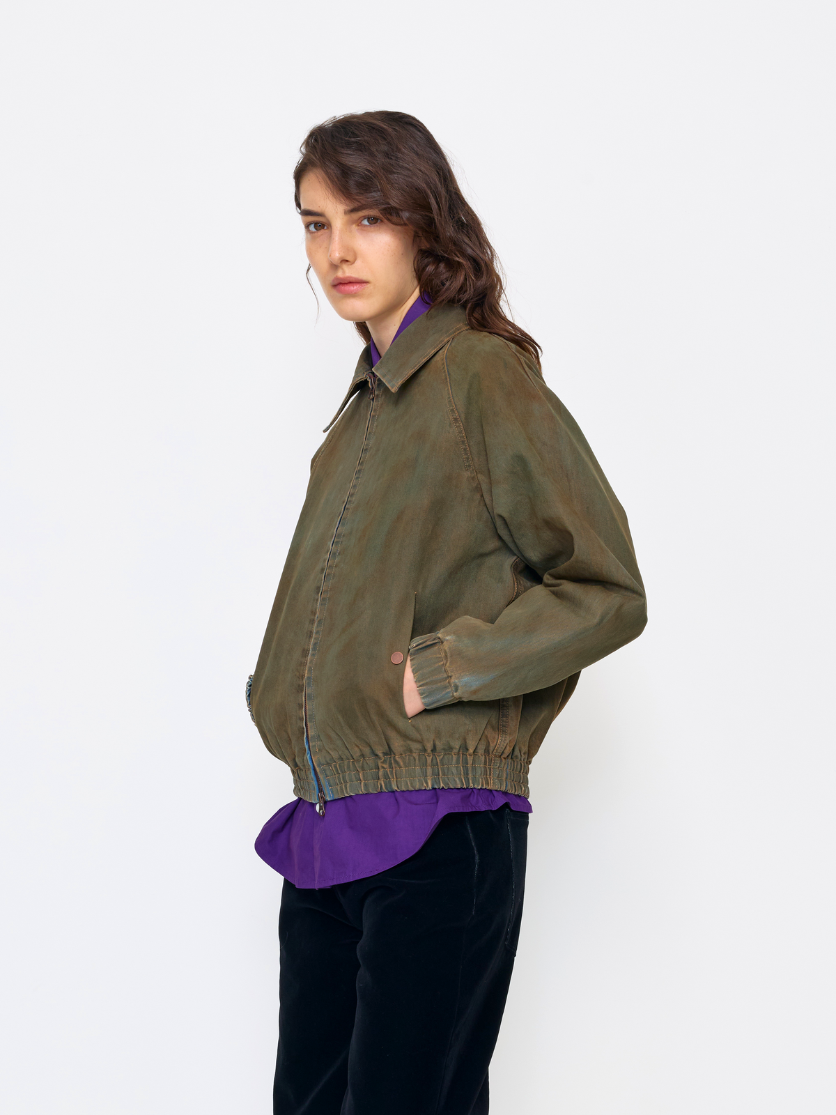 A model wearing an olive green utility-style jacket over a purple collared shirt and black pants, photographed against a plain light gray studio background.